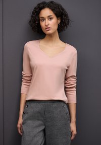 Helles rosa langärmliges Oberteil mit V-Ausschnitt, kombiniert mit grauen karierten Hosen, die seitliche Knopfakzente aufweisen. Strukturierter Stoff.