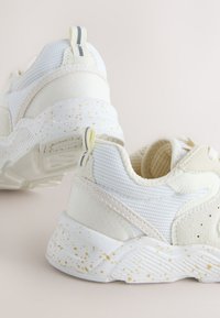 Baskets blanches en mesh et suède avec une semelle intermédiaire beige mouchetée et des languettes, inclinées pour montrer les détails du talon et du côté intérieur.