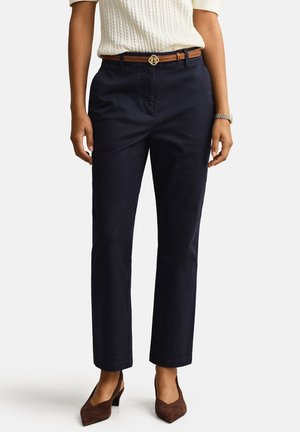 Vrouw die marineblauwe rechte broek draagt, met een bruine riem met een gouden gesp, een witte trui, bruine pumps met spitse neus en een polshorloge.