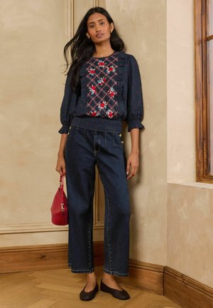Frau mit dunkler Blumenbluse und weit geschnittenen Jeans hält eine rote Handtasche und steht drinnen nahe dem Fenster.