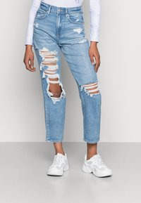 Jean en denim délavé clair avec des déchirures usées, taille haute, coupe droite et ourlet brut. Associé à des baskets athlétiques blanches.