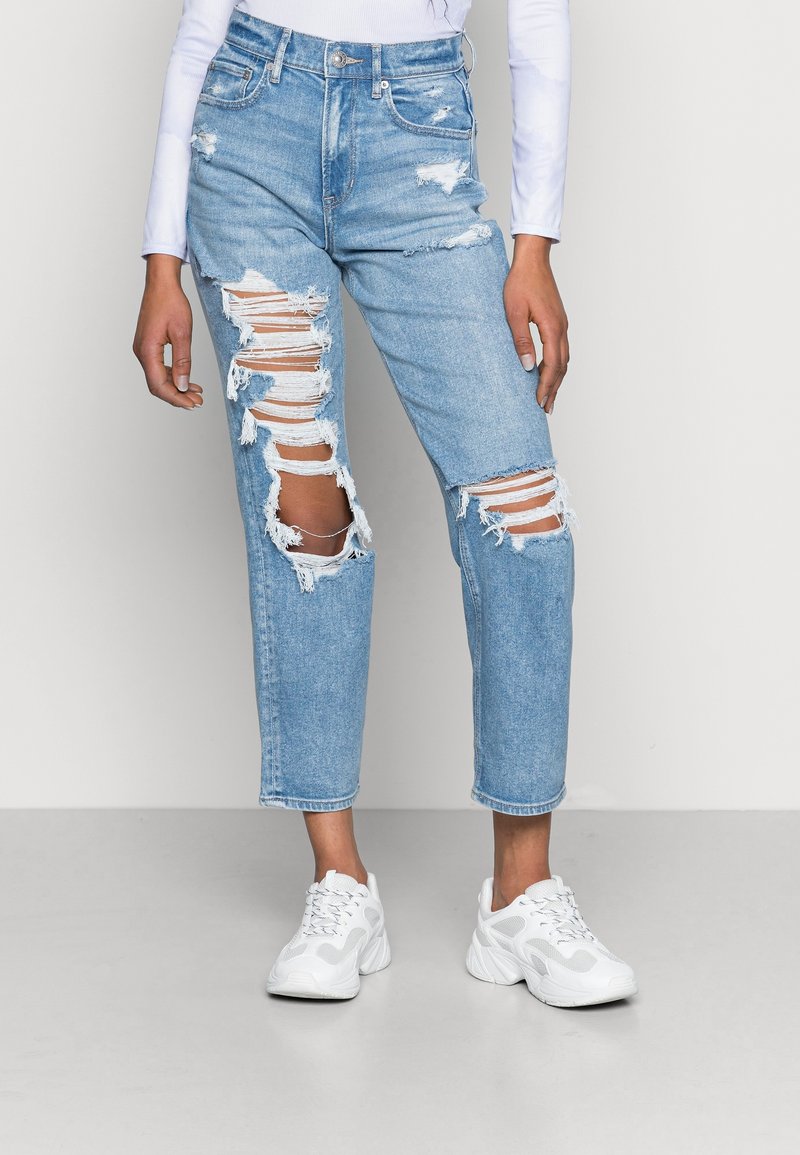 Jean en denim délavé clair avec des déchirures usées, taille haute, coupe droite et ourlet brut. Associé à des baskets athlétiques blanches.