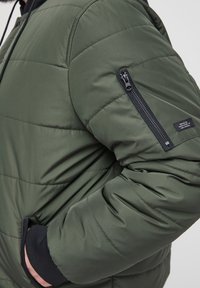 Grønn boblejakke med horisontal quilting, glidelåslomme på venstre arm, elastiske mansjetter og en jevn, blank tekstur.