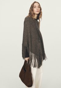 Poncho lavorato a maglia di colore marrone con bordi sfrangiati, indossato sopra una maglietta nera. La modella tiene in mano una borsa in pelle scamosciata marrone scuro. Sono visibili pantaloni di colore chiaro.