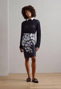 Pull en laine bleu marine avec un col blanc, associé à une jupe noire ajustée ornée de motifs floraux blancs. Le modèle porte des sandales noires à glissière.