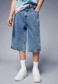 Lys blå denimshorts med avslappet passform, fremre lommer og rå kant, kombinert med hvite sneakers og ankelstrømper.
