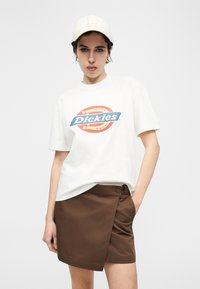 Νεαρός που φοράει λευκό μπλουζάκι με το λογότυπο Dickies, καφέ φούστα τύπου wrap, μπεζ καπέλο μπέιζμπολ με το λογότυπο LA και κρίκους στα αυτιά, ποζάροντας με το ένα χέρι στην τσέπη.