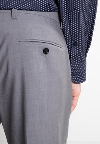Pantalonii gri, croiți pe măsură, au un buzunar la spate cu un buton negru, asortați cu o cămașă albastră cu model, ce prezintă o textură netedă.
