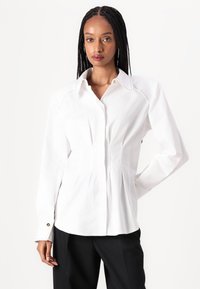 Blouse blanche à manches longues avec un col pointu, fermeture à boutons, devant plissé et coutures décoratives aux épaules.