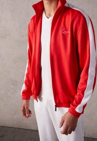 Chaqueta roja con cremallera y rayas laterales blancas, con un cuello alto y puños elásticos. Lleva puesta una camiseta blanca de cuello en V, de textura suave.