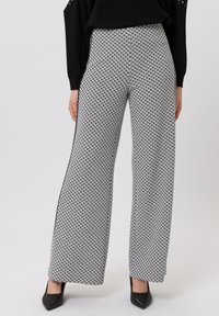 Wijd-uitlopende broek met een zwart-wit klein geometrisch patroon, zwarte zijstreepjes, gedragen met zwarte puntige hakken en een zwart topje.