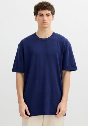 Joven de pie inmóvil vistiendo una camiseta de manga corta suelta de color azul oscuro y pantalones beige claros sobre un fondo liso y claro.