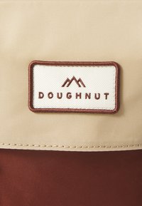 Tissu beige avec une section inférieure bordeaux, doté d'un label blanc portant la mention "DOUGHNUT" avec des icônes de montagnes marron au-dessus du texte.