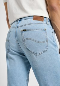 Světle modré džíny s zadními kapsami a viditelným prošitím, nošené osobou v bílém tričku na jednobarevném pozadí.