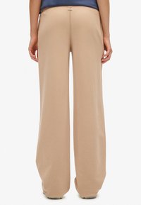 Beżowe spodnie o szerokich nogawkach wykonane z miękkiej tkaniny, charakteryzujące się gładką fakturą i subtelnym detalem szwów. Widok od tyłu pokazuje prosty pas z małą metką.