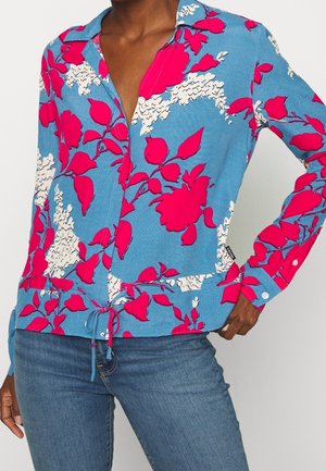 Kvinde iført blå skjorte med knapper og et lyst pink og hvidt blomsterprint, kombineret med blå jeans, hånd på hoften, stående foran hvid baggrund.