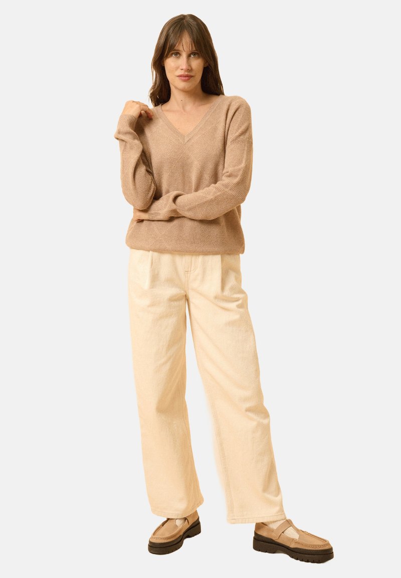 Beige stickad tröja med V-ringning, parat med lösa krämfärgade byxor och ljusbruna skor med mörka yttersulor. Slät textur, avslappnad design.