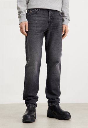 Osoba ubrana w wyblakłe czarne dżinsy i czarne masywne botki stojąca na jasnej podłodze na jasnym tle.