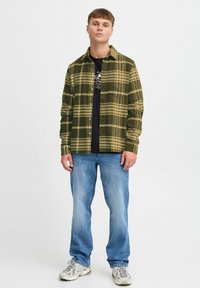 Grøn og gul ternet flannel skjorte med knaplukning fortil, båret over en sort t-shirt, parret med lyseblå jeans og grå sneakers.
