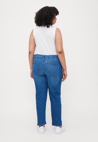 Jeans de ganga azuis com corte reto, apresentando uma cintura média, bolsos traseiros e uma bainha virada; combinados com sapatos brancos.