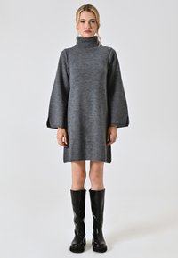 Abito grigio lavorato a maglia con collo alto e maniche lunghe a campana. Abbinato a stivali neri fino al ginocchio e una silhouette semplice.