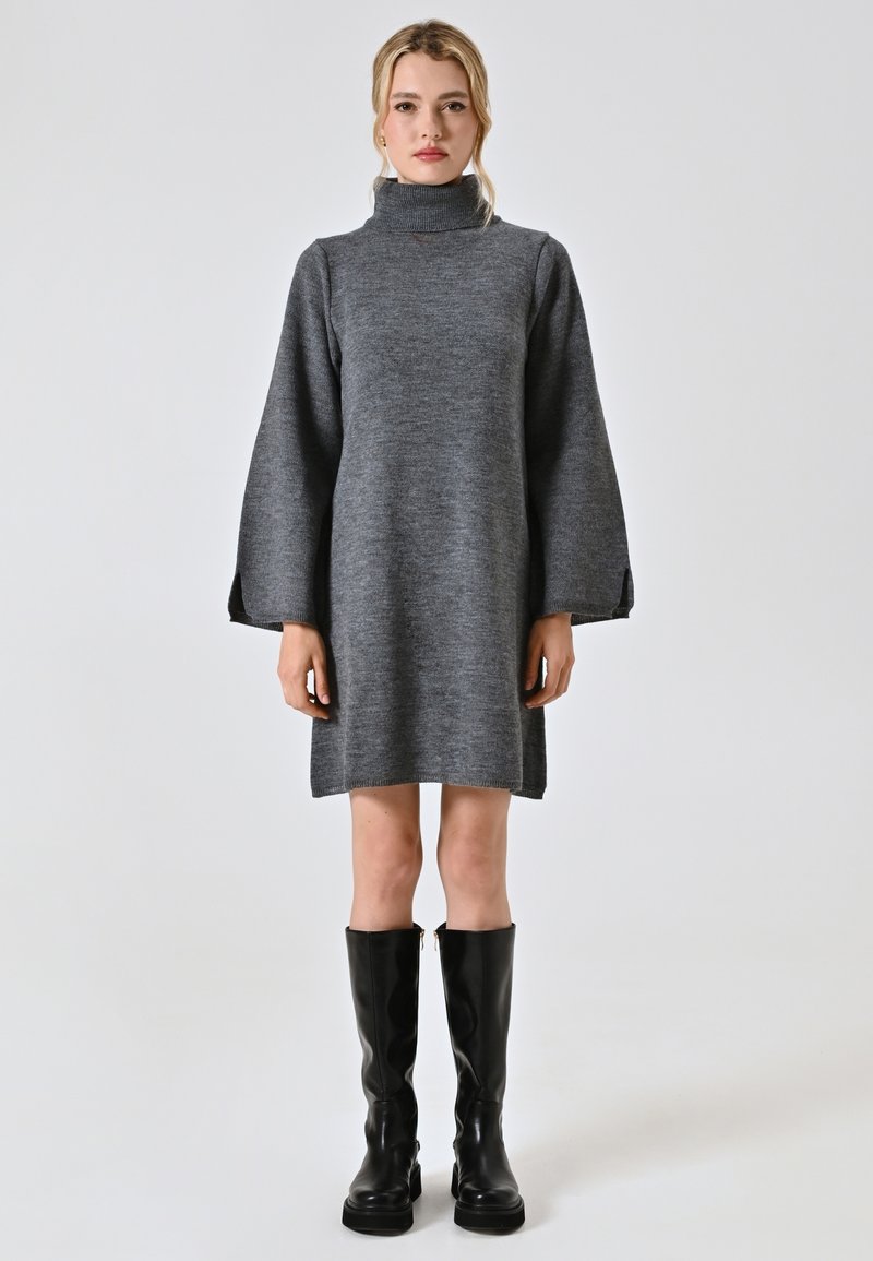 Abito grigio lavorato a maglia con collo alto e maniche lunghe a campana. Abbinato a stivali neri fino al ginocchio e una silhouette semplice.