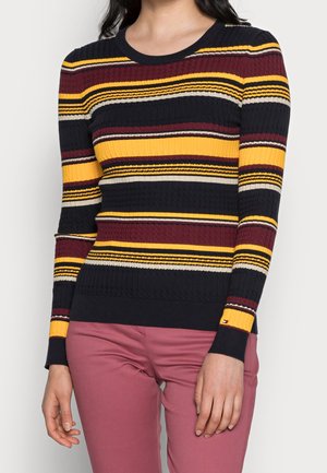 Femme portant un pull côtelé à manches longues avec des rayures horizontales jaunes, noires, bordeaux et blanches et un pantalon rose.