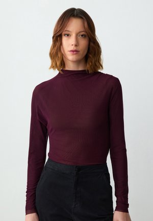 Maglietta lunga color marrone bordeaux con collo alto, texture morbida e design aderente, abbinata a pantaloni neri a vita alta.