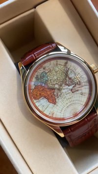 Ceas de mână cu nuanță de aur, având un cadran cu hartă, care dispune de o brățară din piele texturată de culoare maronie. Geamul este clar, evidențiind detaliile geografice.