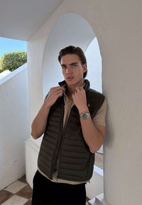 Młody mężczyzna poprawiający kołnierz ciemnej kamizelki na beżowej koszulce polo, stojący pod białą arkadą z widoczną zieleń na zewnątrz.