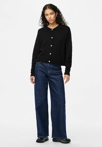 Črni kardigan z ovratnikom in zapiranjem spredaj na gumbe, kombiniran z razširjenimi temno modrimi kavbojkami in črnimi čevlji. Tkanina z teksturo.