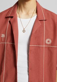 Camicia color ruggine con maniche corte, caratterizzata da ricami bianchi di palme e soli, e cuciture decorative lungo le cuciture.