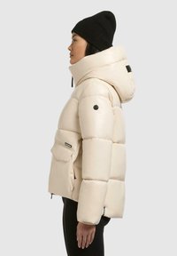Giacca puffer beige con cappuccio oversize e tasca frontale, texture liscia, finitura lucida, accentuata da una patch del logo nera e zip.