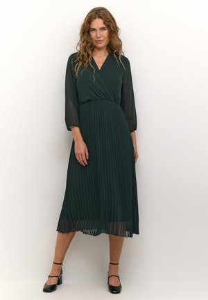 Green pleated midi dress with a wrap bodice and sheer long sleeves, complemented by ankle-strap black heels and a flowing silhouette.