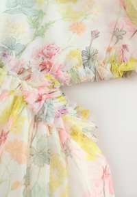 Priesvitná látka s kvetinovým vzorom v pastelových farbách, ozdobená volánikmi a viditeľnými záhybmi, vytvárajúca jemný a textúrovaný vzhľad.