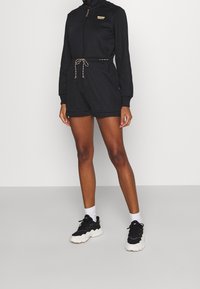 Černý sportovní overal s dlouhými rukávy, zipem a stahovacími šňůrkami v pase. Doplněný černými teniskami a bílými kotníkovými ponožkami.