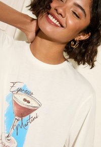 Valkoinen puuvillainen T-paita, jossa on graafinen kuva espresso martini -juomasta. Kuvassa on ruskeita ja kermansävyisiä värejä, sekä vaaleansinisiä koristeita lasin taustalla.