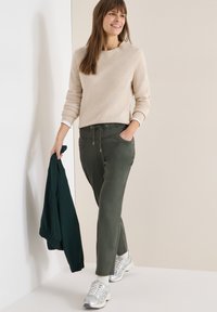 Femme souriante, portant un pull beige, un pantalon vert à cordon, des chaussettes blanches, des baskets argentées, tenant une veste vert foncé dans la main gauche, debout à l'intérieur.