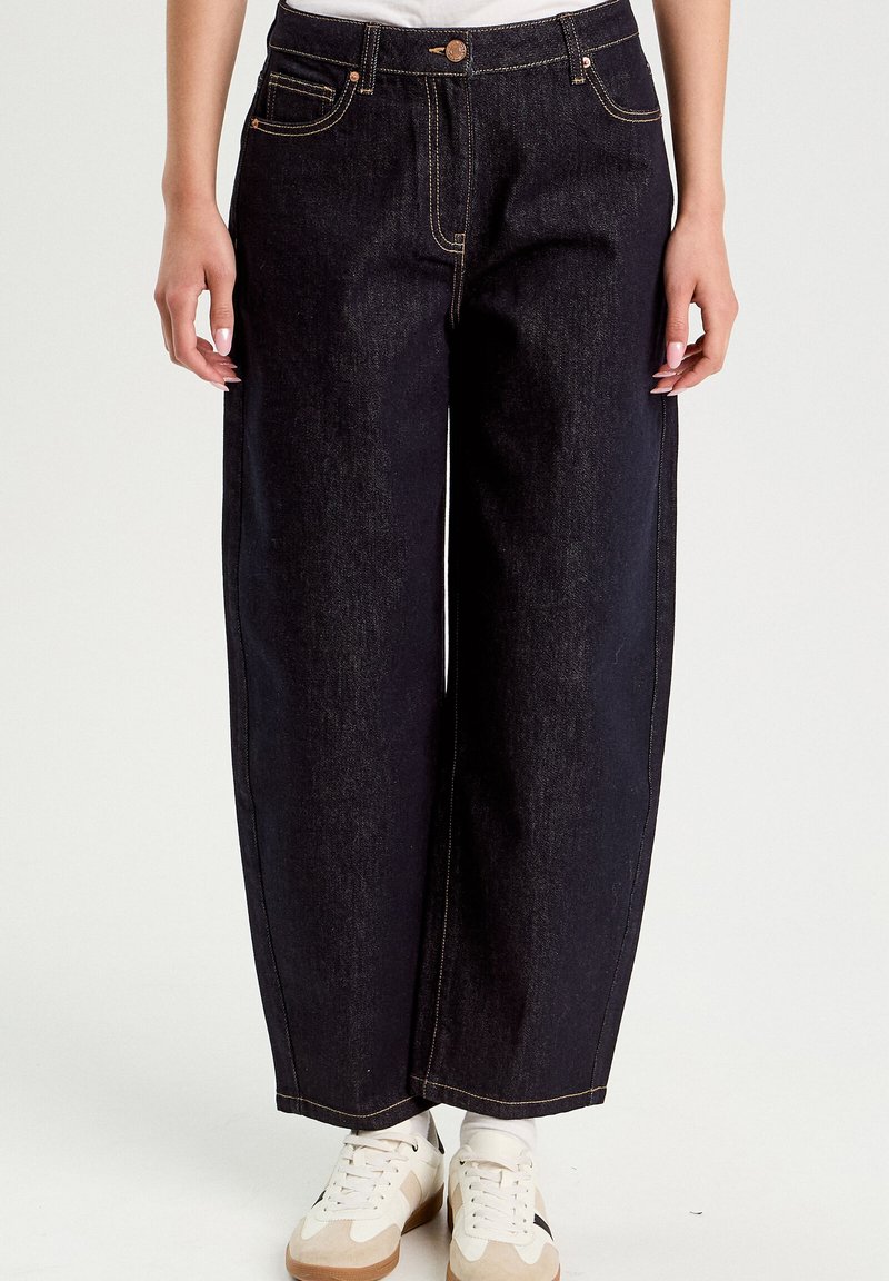 Jeans in denim scuro a gamba dritta con cuciture a contrasto, indossati con sneakers bianche e calzini bianchi.