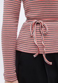 Κοντινό πλάνο ενός μακρυμάνικου τοπ με κόκκινη, λευκή και μαύρη ρίγα, με ζωνάκι δεμένο στη μέση, συνδυασμένο με μαύρο παντελόνι.