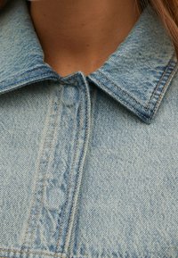 Close-up van de kraag van een lichtblauwe denimjas met zichtbare stiksels en een gedeeltelijk dichtgeknoopte voorkant, gedragen door een persoon met een lichte huid.