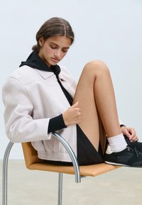 Veste bomber rose clair avec une capuche noire, assise sur une chaise moderne. Une robe noire courte, des chaussettes blanches et des baskets noires complètent la tenue.