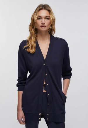 Femme aux longs cheveux blonds ondulés, portant un cardigan bleu marine à boutons avec des accents dorés et un pantalon assorti, se tenant devant un fond uni.