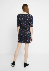 Młoda kobieta z długimi brązowymi włosami, ubrana w czarną krótką sukienkę w kolorowe abstrakcyjne wzory, czarne sneakersy za kostkę, stoi tyłem.