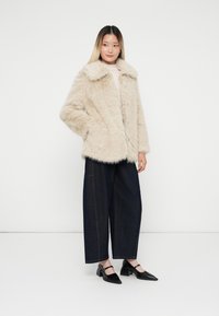 Giacca in pelliccia sintetica beige con colletto ampio, abbinata a pantaloni in denim scuri larghi e scarpe nere con tacco. Adatta per un look casual.