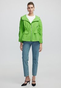 Groene lichtgewicht jas met een capuchon, cinched taille en twee voorzakken. Gecombineerd met lichtblauwe jeans en zwarte haksandalen.