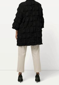 Black textured oversized jacket with a quilted pattern, three-quarter sleeves, paired with cream-colored trousers and black high-heeled shoes.
