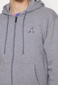 Personne portant un sweat à capuche gris zippé avec un logo Jumpman noir et blanc sur le côté gauche de la poitrine et des cordons suspendus.