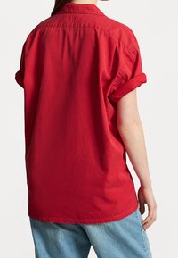 Person wearing loose red short-sleeve shirt with rolled cuffs and light blue jeans, shown from the back against a plain background.