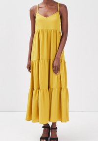 Robe maxi jaune à volants avec bretelles tressées, encolure en V et silhouette fluide. Mise en valeur par un ourlet évasé et un tissu doux.